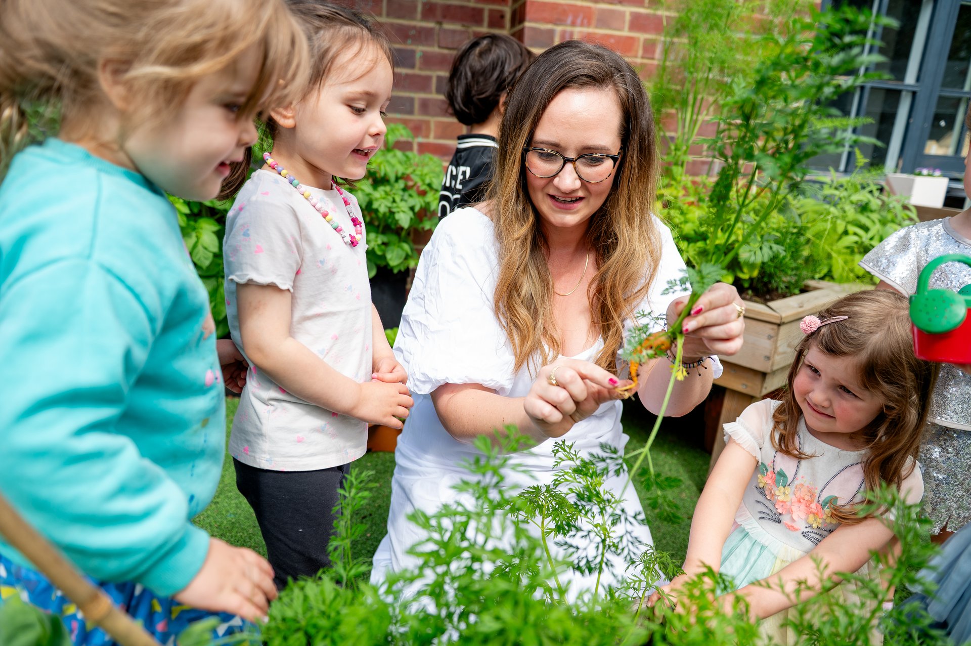 A smiling teacher helps three children explore plants in a sunny school garden, holding a sprig as they gather around raised beds.