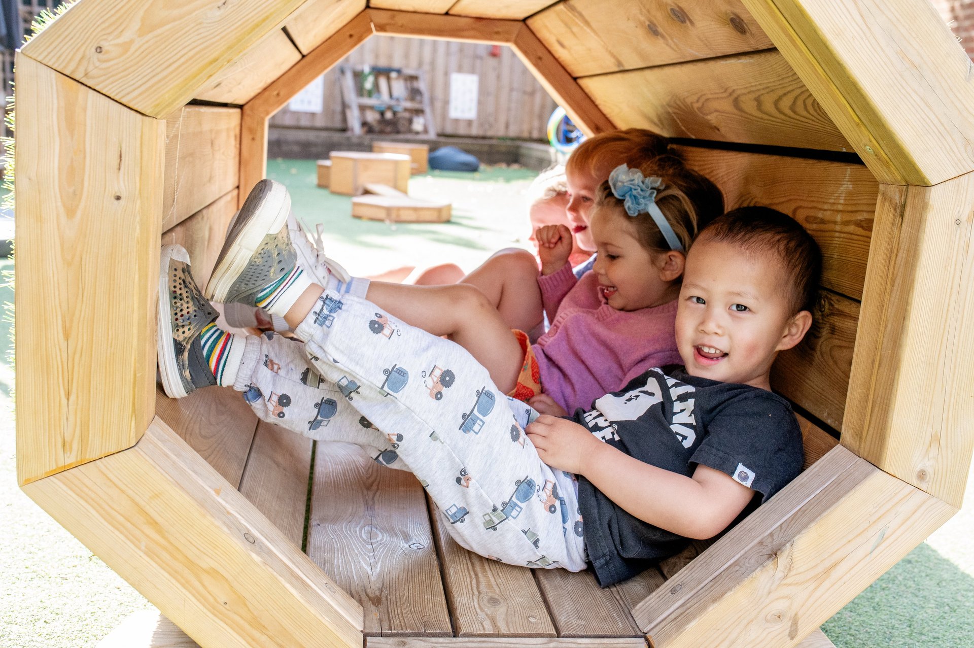 Three cheerful children relax together inside a hexagonal wooden playhouse, smiling and playing in a cozy, sunlit outdoor playground.
