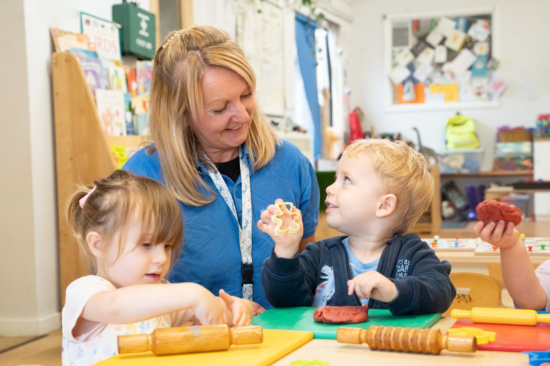 Teacher helps two young children bake at a classroom table; boy holds dough, girl rolls pastry, creating a warm, joyful learning scene.